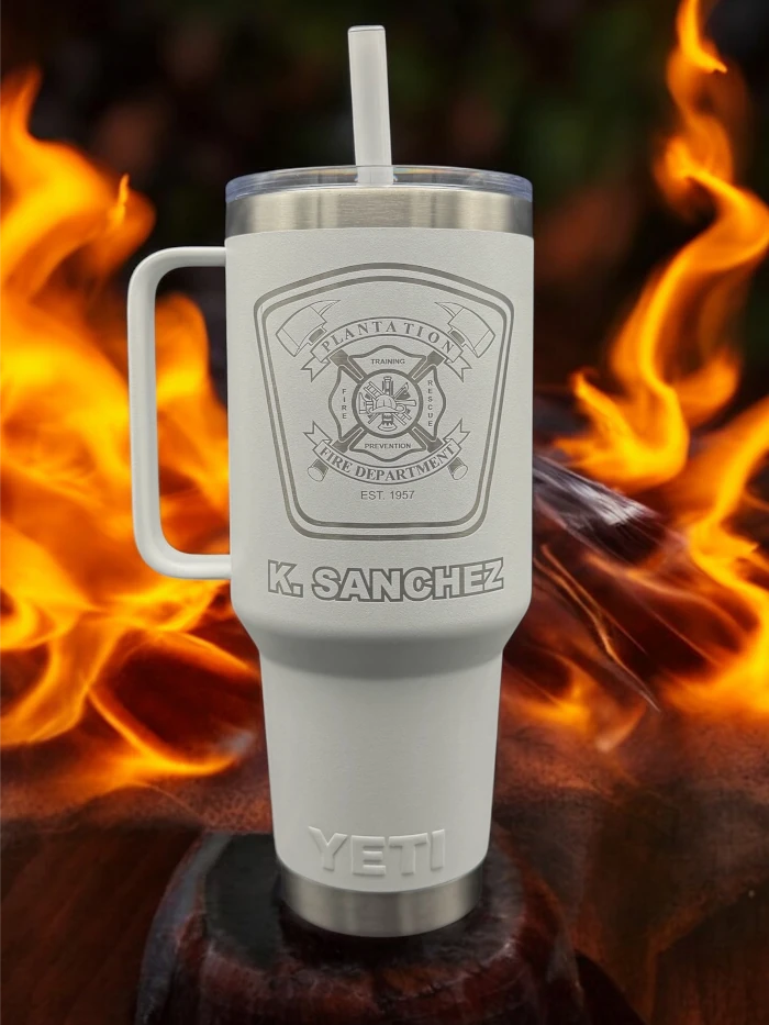 A white Yeti tumbler with a straw, engraved with a fire department badge and the name "K. Sanchez," is placed in front of flames.