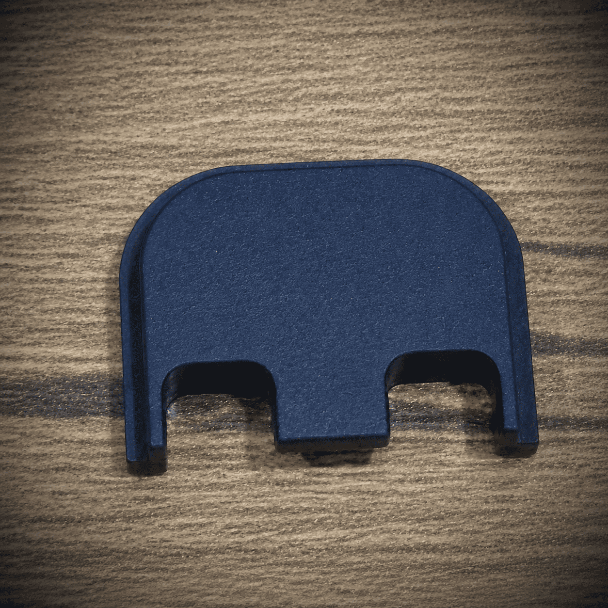 A black plastic or metal component with two prongs sits on a wood grain surface.