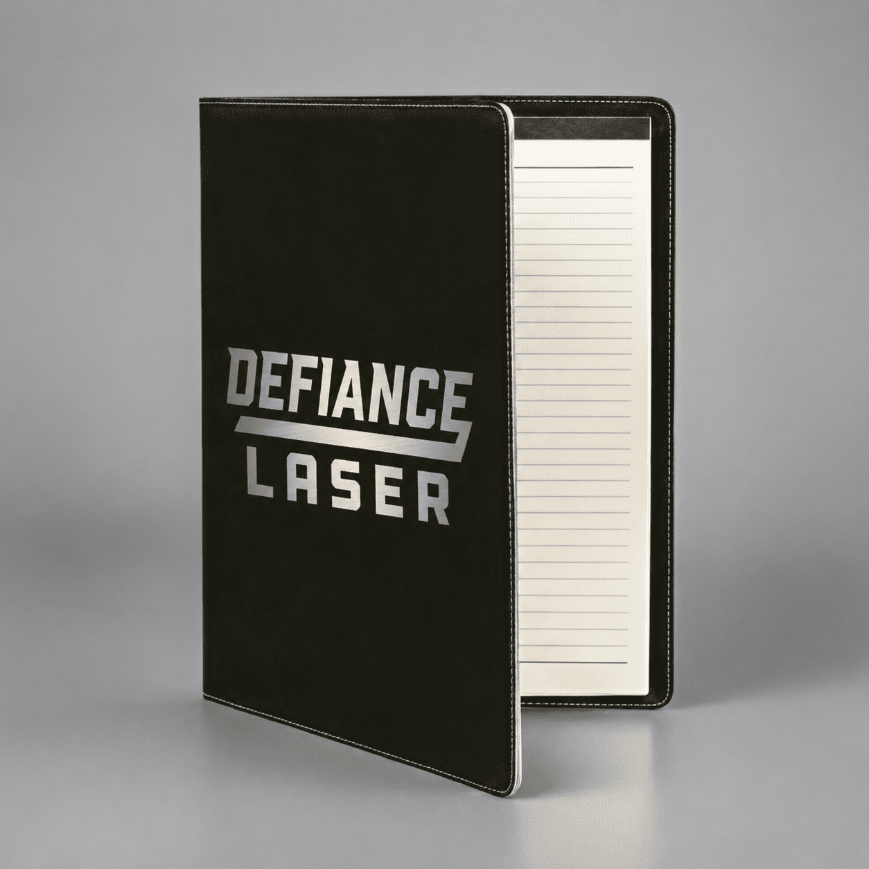 A black portfolio folder with "DEFIANCE LASER" printed in bold silver letters on the cover, shown standing open with lined paper inside.